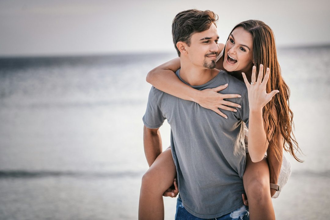 A smiling woman on a man's back at the beach, showing her engagement ring