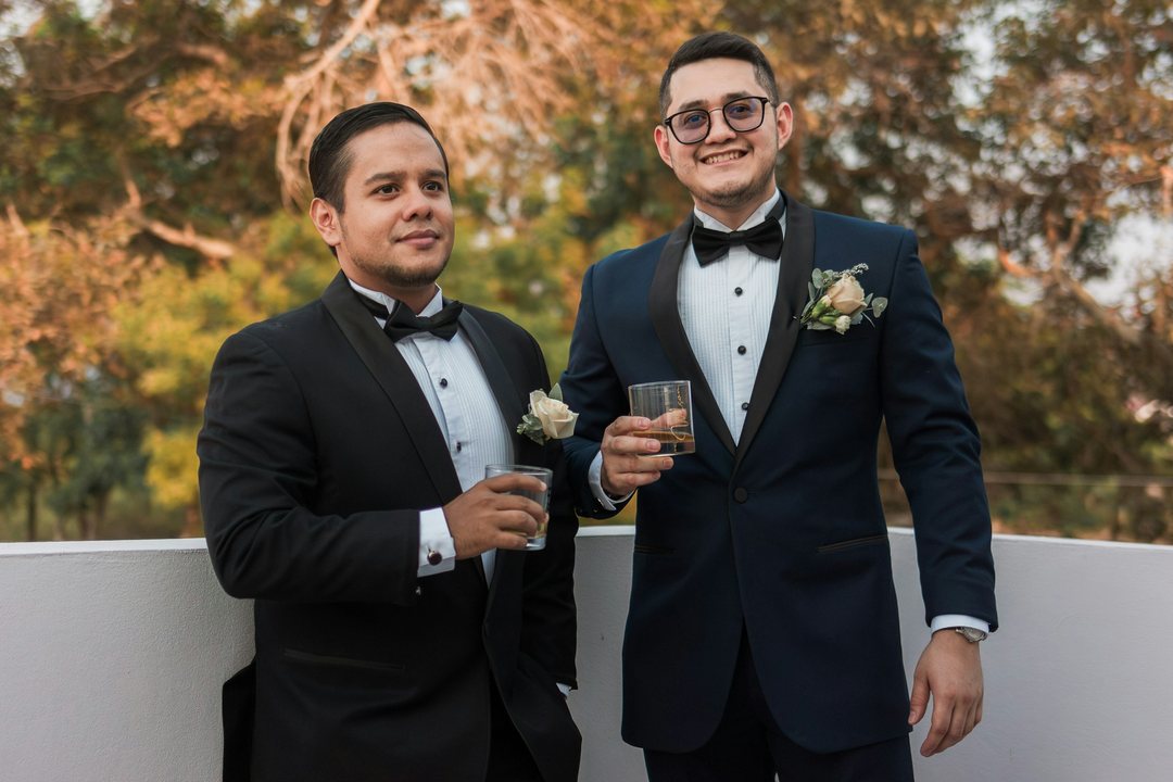 Two smiling grooms in tuxedos and bow ties holding drinks outdoors